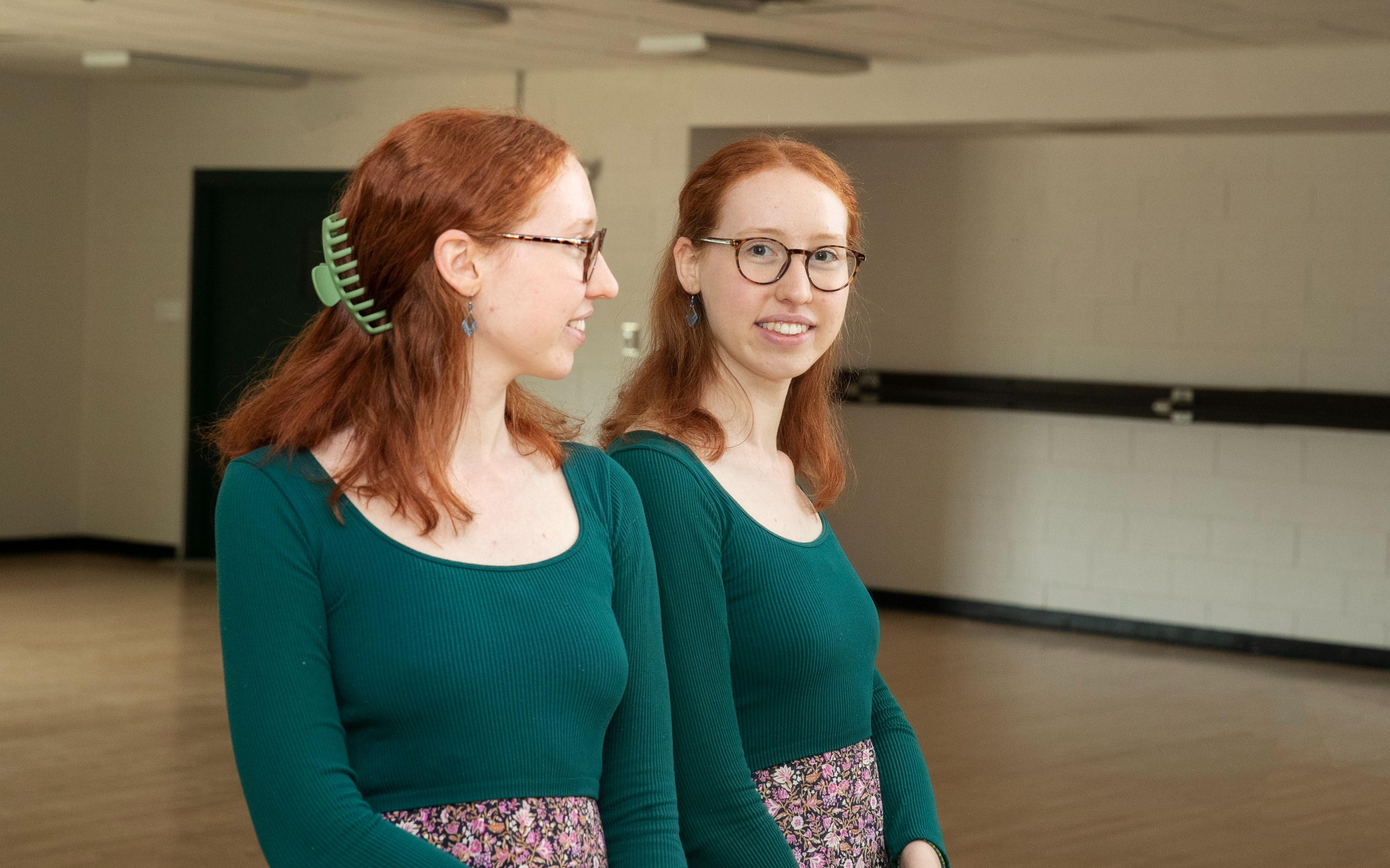 Liz Bracht '24 smiles into a mirror in a dance studio. She is wearing a blue, long sleeved top and a floral skirt.
