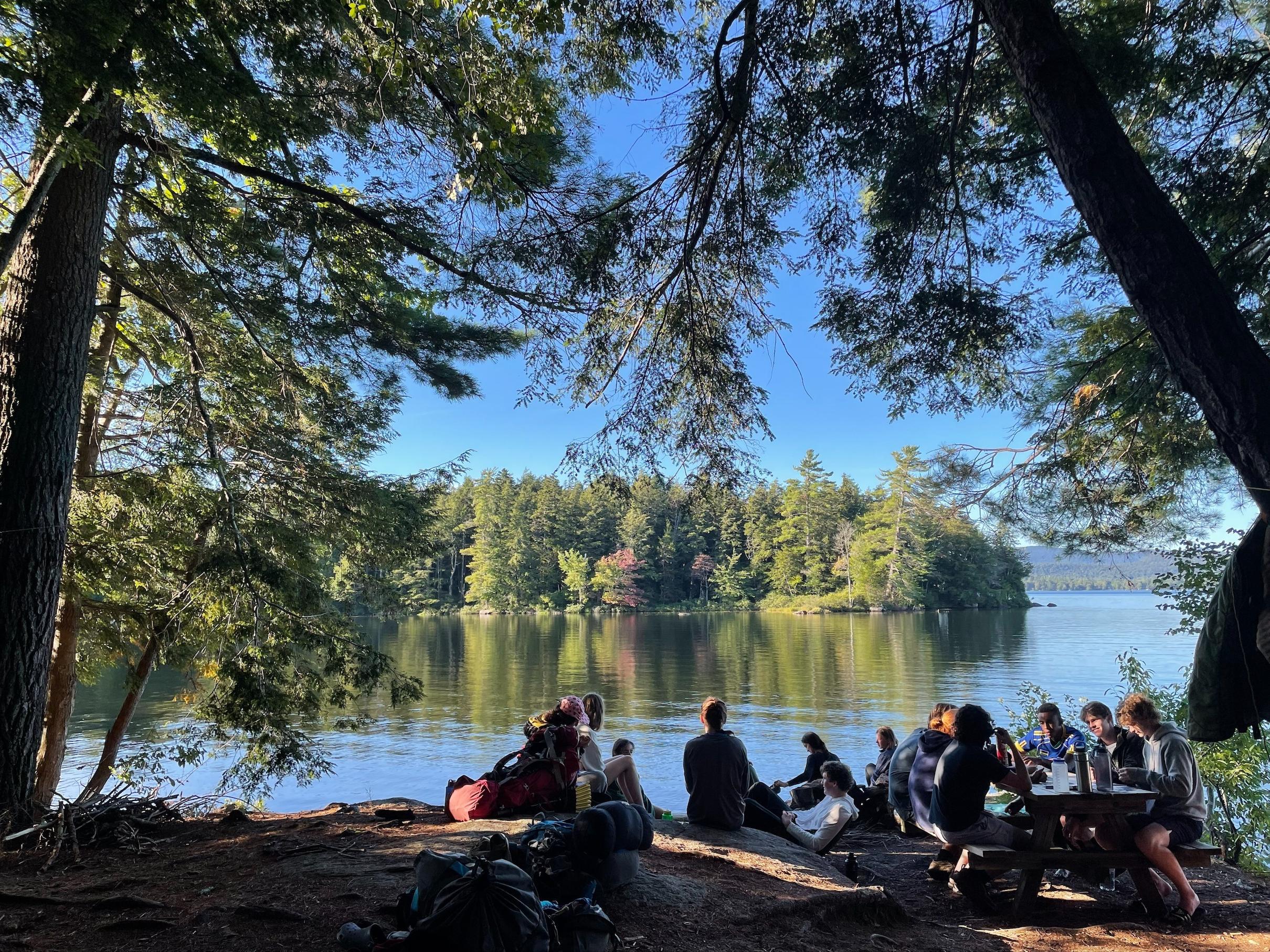 We see the backs of a group of students as they chat at a picnic table and look out across a lake.