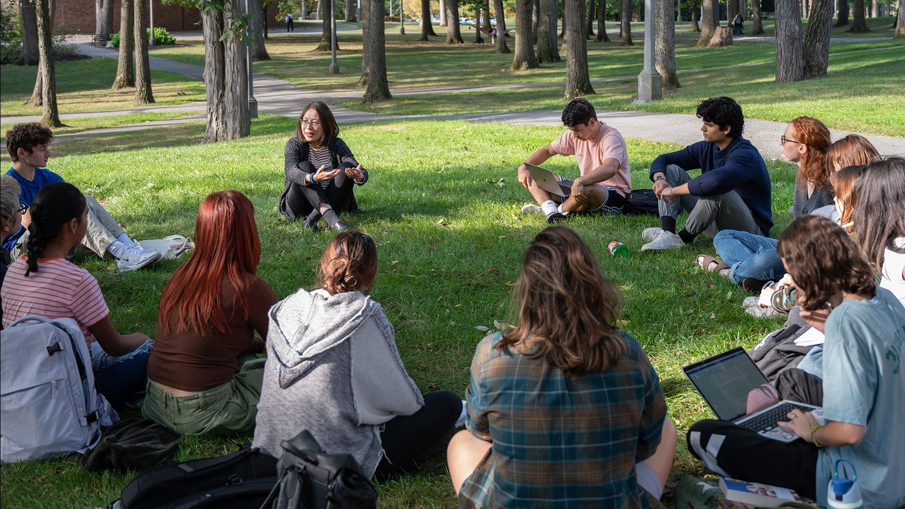 Professor Mao Chen sits in a circle on the Skidmore green with her Ideal Worlds students. They listen, engaged, as she speaks.